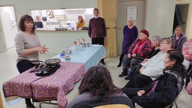 Woman speaking to a group in front of cooking supplies