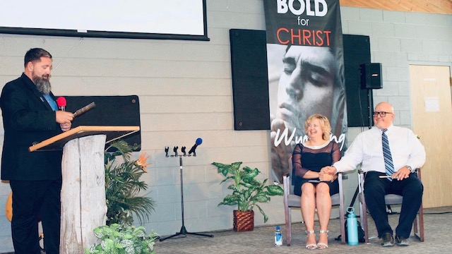 Man speaking at a podium and a pastor and his wife sitting to the side